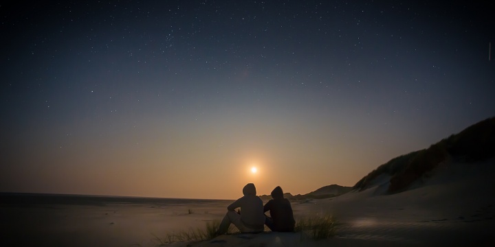 Two people sitting on a beach with the sun setting. 