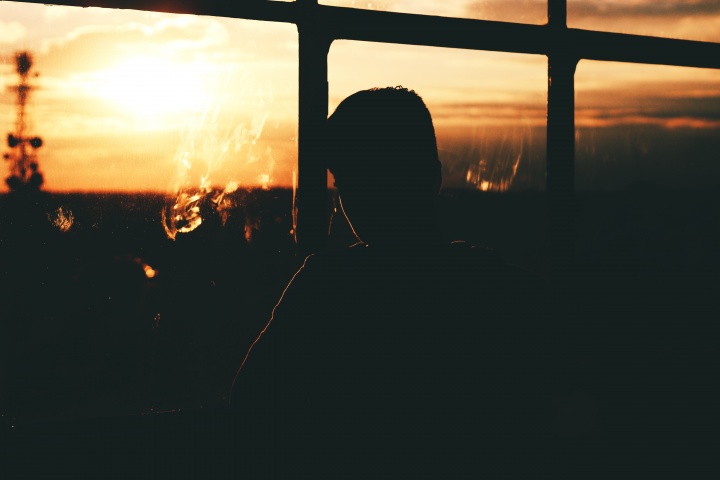 A young man looking out a window with the sun setting.