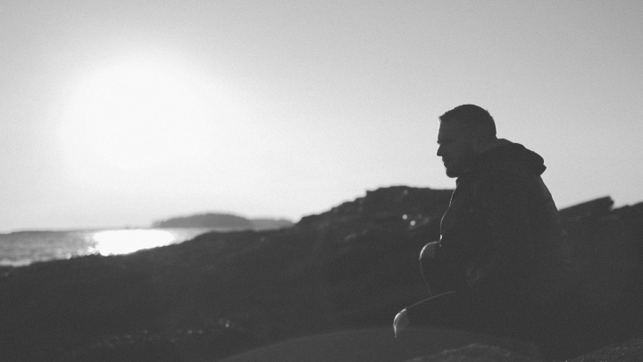 A young man sitting looking out at water.