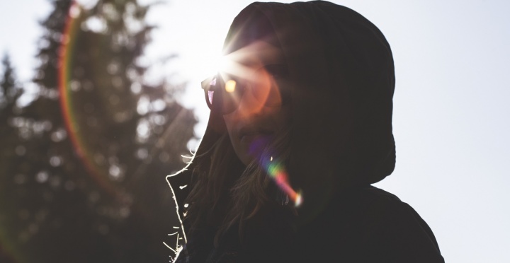 A girl wearing a jacket and hood looking at the camera with sunrays behind in head.