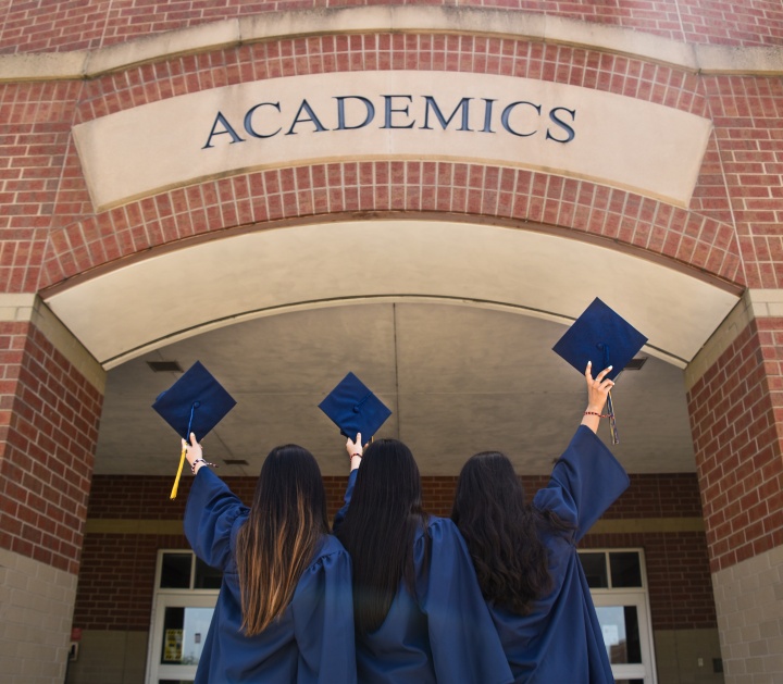 three women in blue graduation robes and holding graduation caps under a brick archway