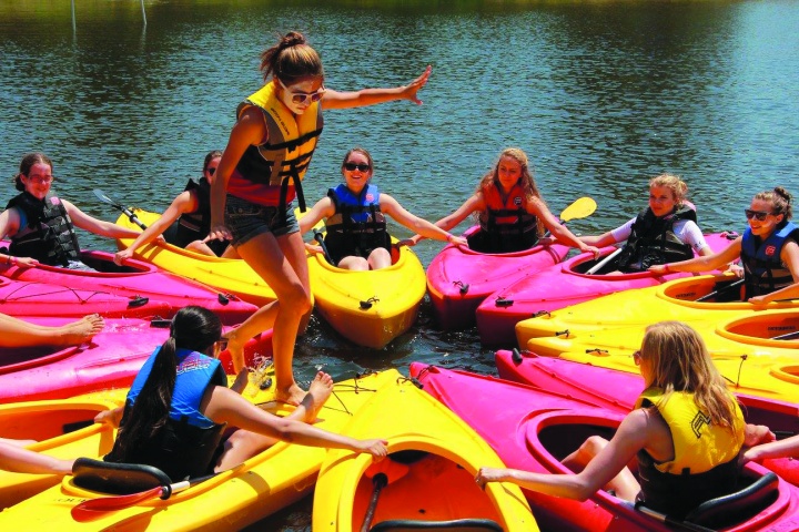 A picture of a group of campers in kayaks form a circle with the kayaks while one camper walks around the circle. 