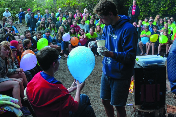 A photo of a camper holding a balloon while another camper paints it with shaving cream. The whole camp is watching in background. 