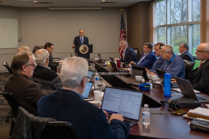 a group of men seated around a table watching a presentation by a man at a lectern