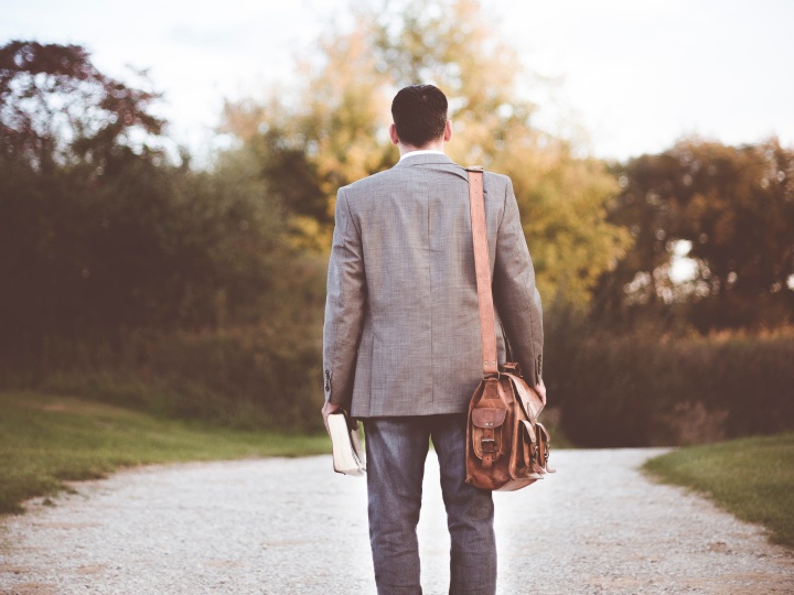 a man dressed in a suit, holding a Bible and with a satchel hanging from one shoulder, standing on a road